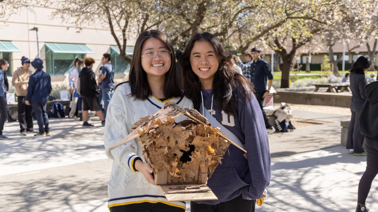 Two smiling students holding a wooden architectural model beneath flowering trees