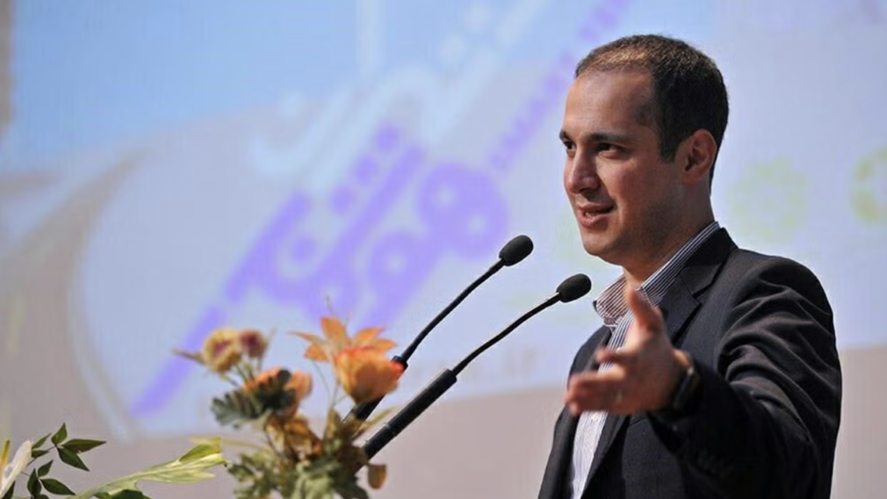 Kaveh Madani in suit speaking at podium with microphones and floral arrangement
