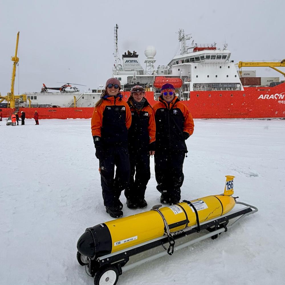 Three people in winter gear stand on snow with a yellow submarine model and an icebreaker ship in the background.