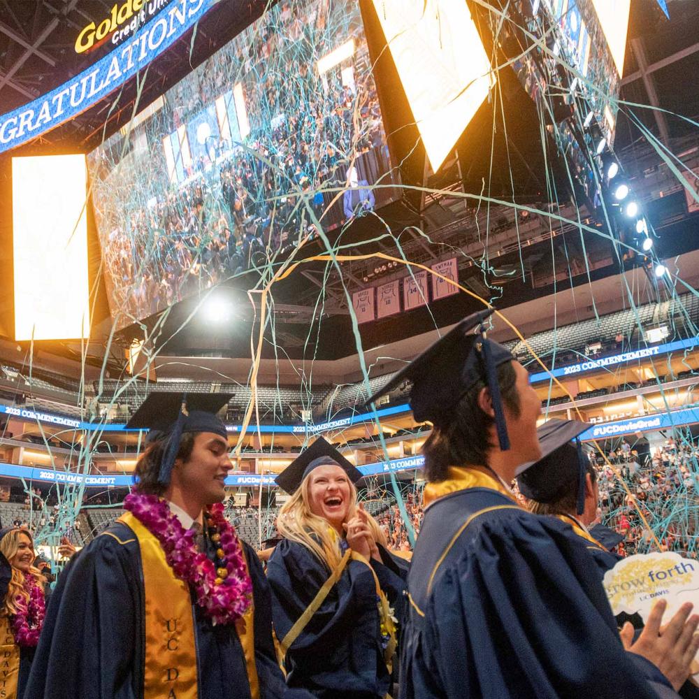 Graduates in caps and gowns celebrating with confetti in an arena under bright lights co