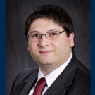 Studio headshot photo of a Barbato in suit and glasses against a dark backdrop