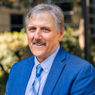Photo of white man with gray hair and mustache outside wearing blue suit jacket, light blue shirt, and blue striped tie. 