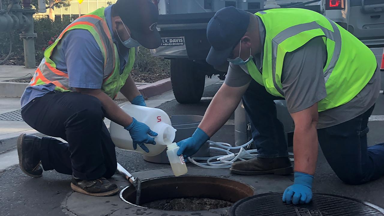 Two workers collecting wastewater samples near an open manhole.