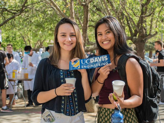 Two female students pose with a smile, holding a College of Engineering sticker