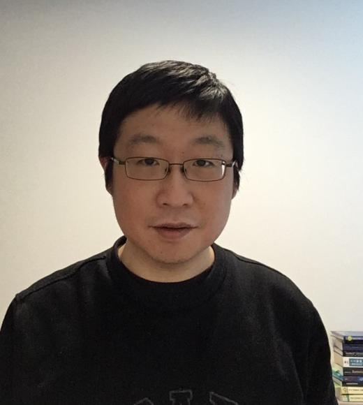 Photo of Chinese man with glasses, short black hair wearing a black shirt against a white wall.