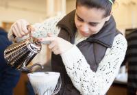 A female student pours coffee from a metal brewing pot into a coffee filter holder.