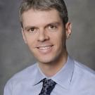 Photo of white man with dark gray hair on gray background wearing light blue shirt with dark blue tie with pattern