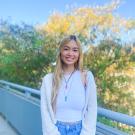 Woman with long blonde hair, tan skin, and brown eyes smiling at the camera. She is wearing a gray cardigan, tank top, and blue jeans. The background is blurry and shows a balcony railing and the leaves of a tree.