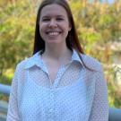 Photo of white woman with dark red hair outside with tree behind her. She is smiling and wearing a white shirt with black polka dots